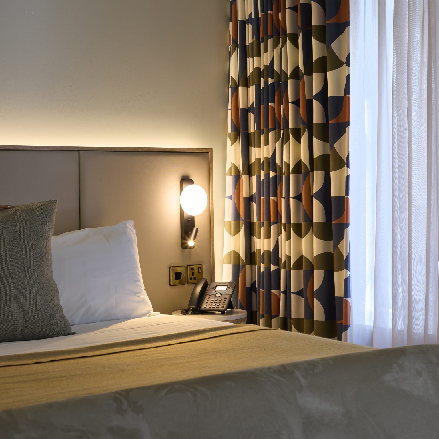 Modern hotel room with stylish upholstered headboard, ambient lighting, geometric curtains, and a bedside phone, creating a comfortable guest experience.