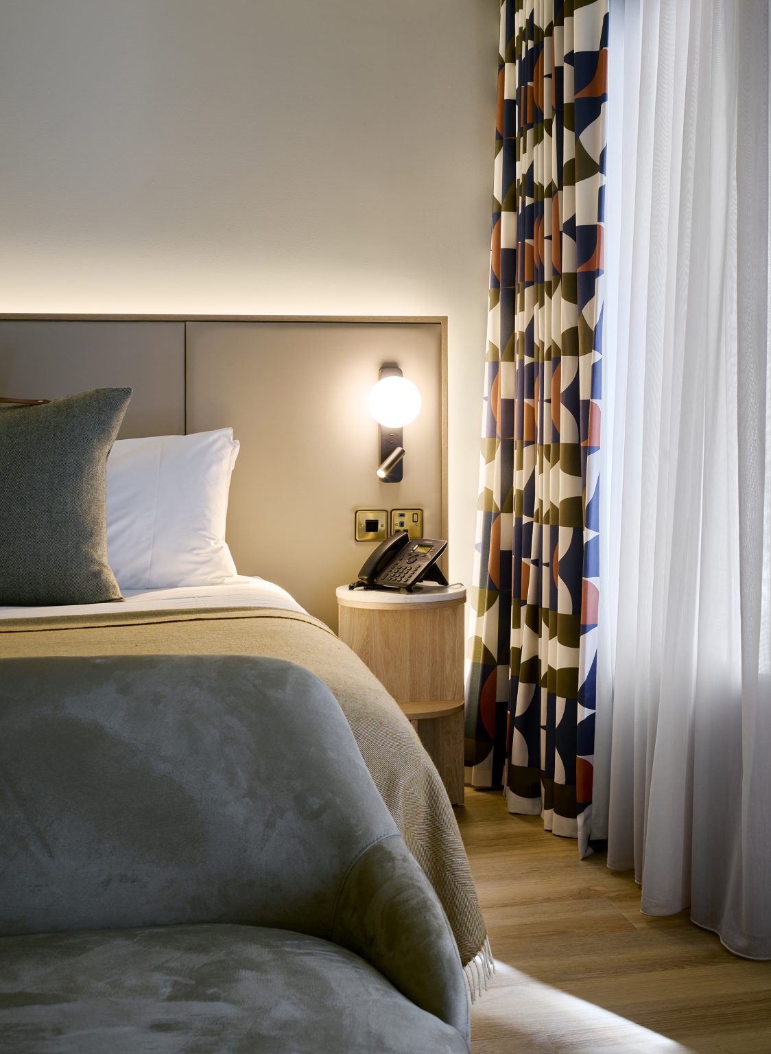 Hotel room with geometric curtains, ambient wall lighting, and a cosy armchair, designed for a relaxing stay in a luxury fit-out.