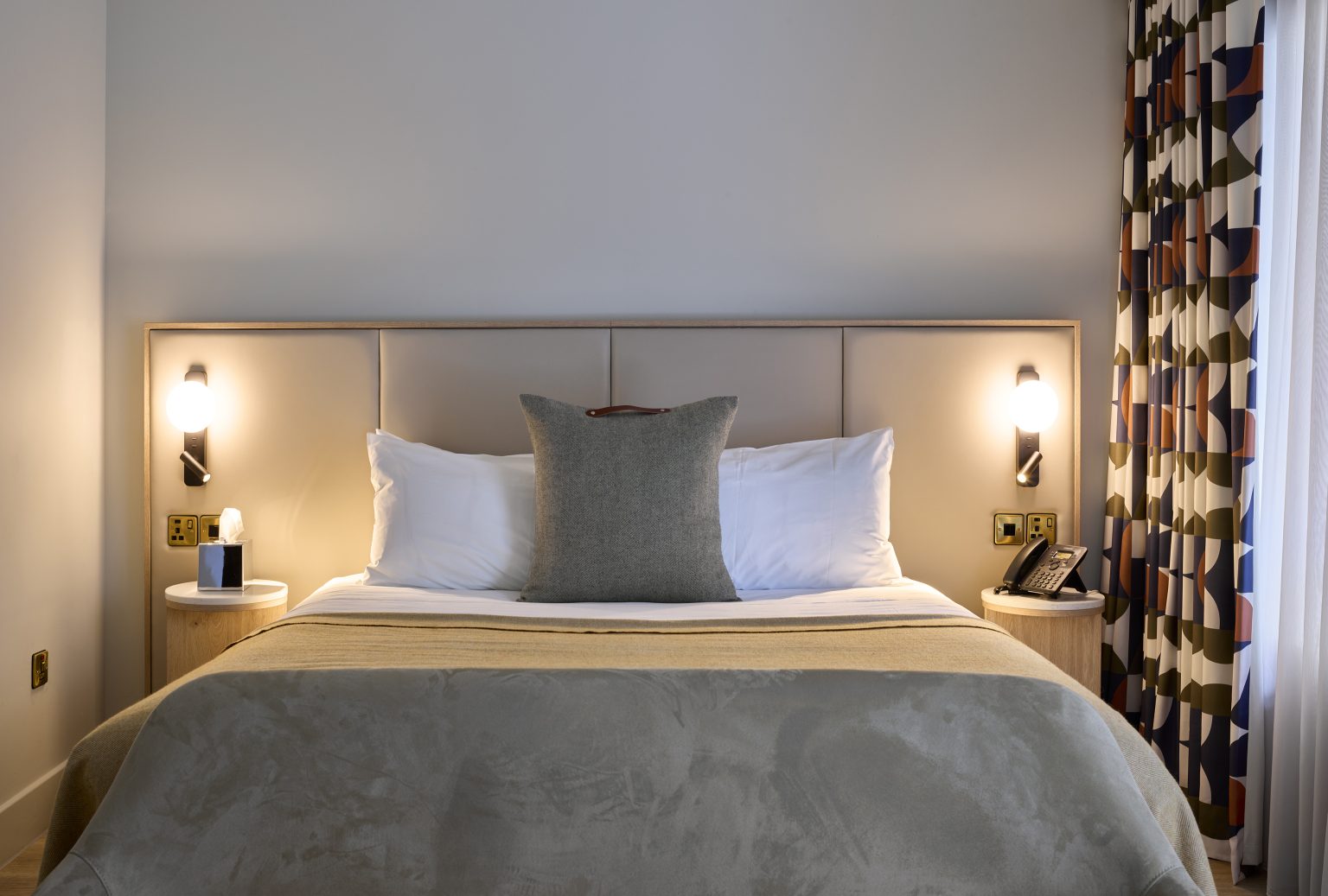 Hotel bedroom with plush pillows, ambient lighting, and geometric patterned curtains, showcasing a modern hospitality fit-out.