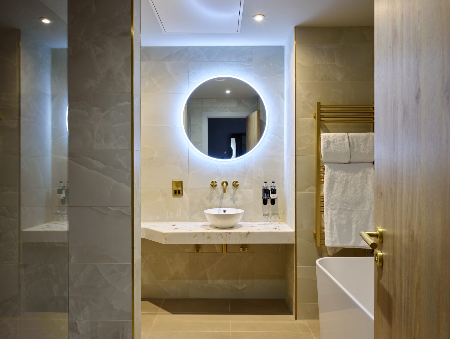 Elegant hotel bathroom featuring a backlit circular mirror, brass fixtures, and a marble vanity for a luxury hospitality experience.