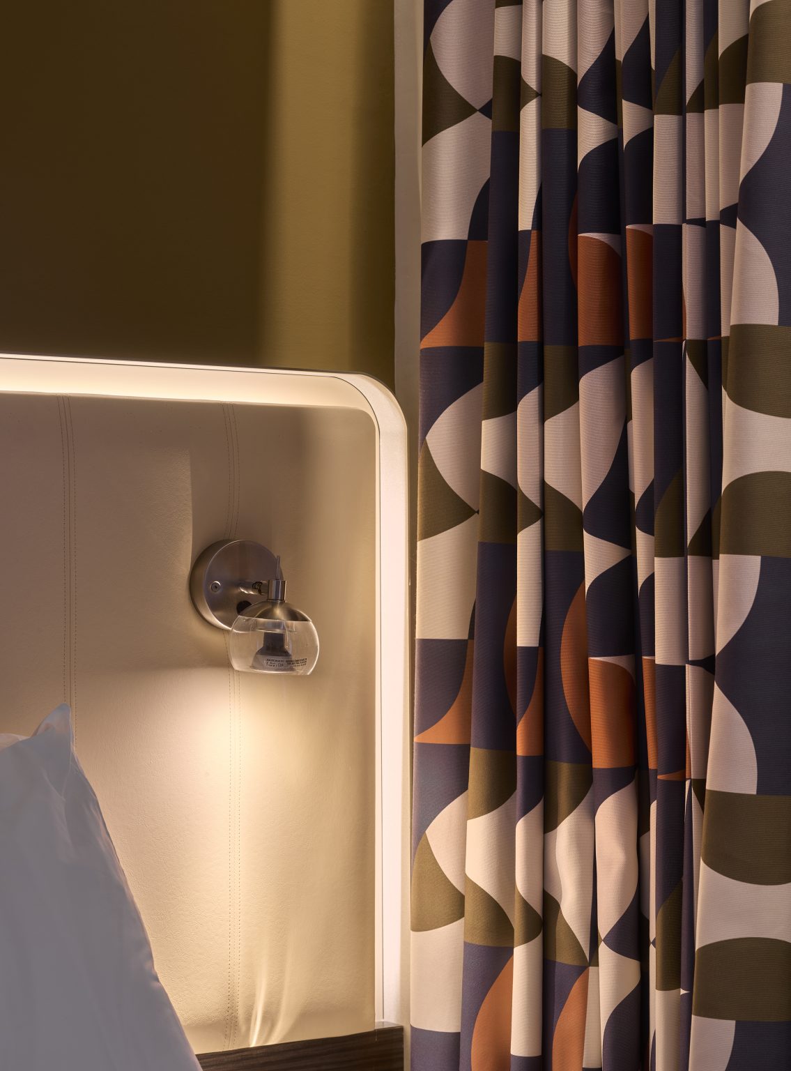 Close-up of stylish hotel room decor with geometric patterned curtains, ambient headboard lighting, and a modern bedside lamp in a hospitality fit-out.