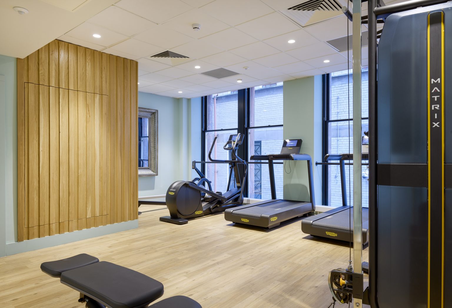 Spacious hospitality gym fit-out with modern exercise equipment, wooden accents, and large windows for natural lighting.