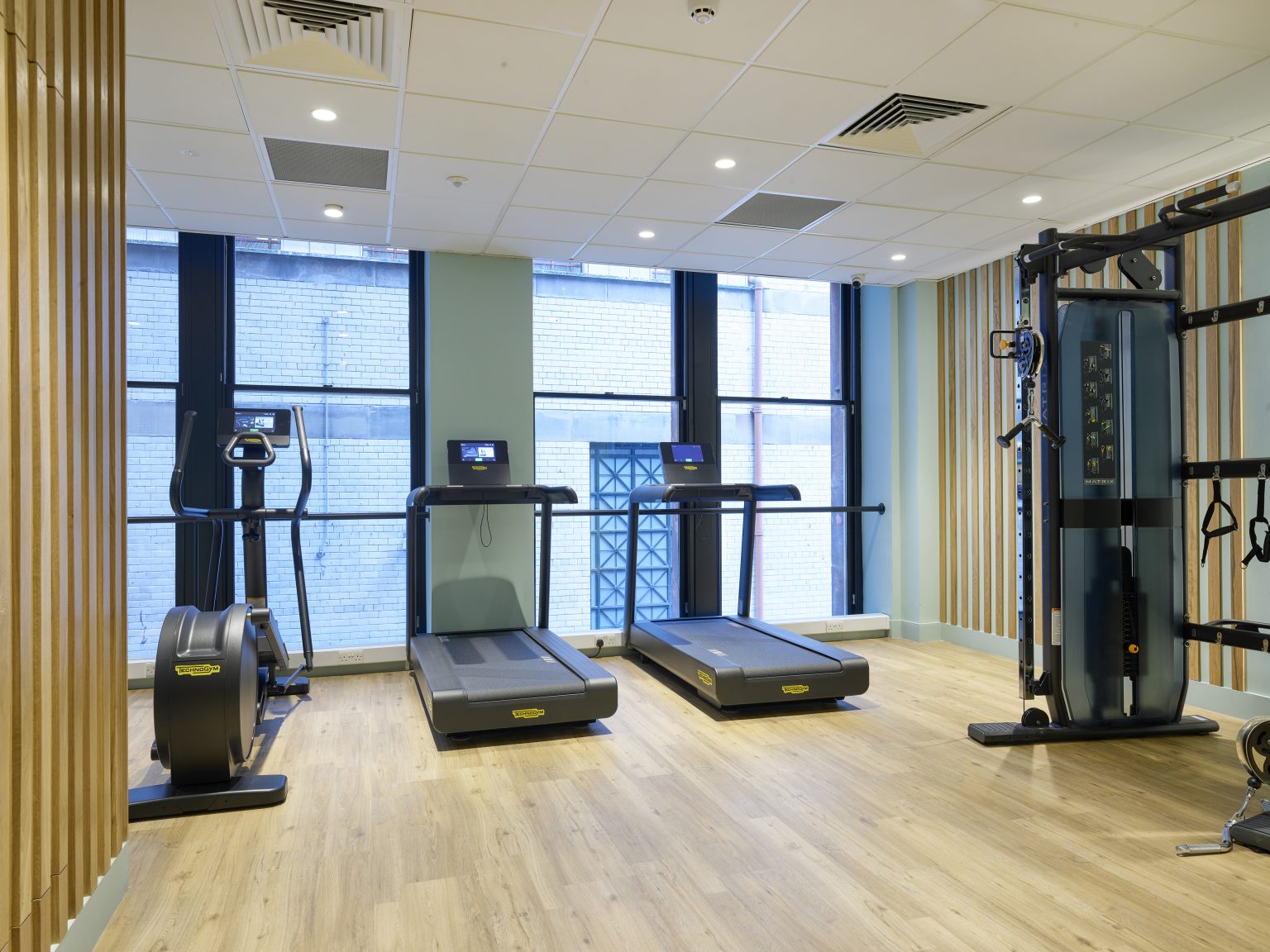 Spacious hospitality gym fit-out with modern exercise equipment, wooden accents, and large windows for natural lighting.