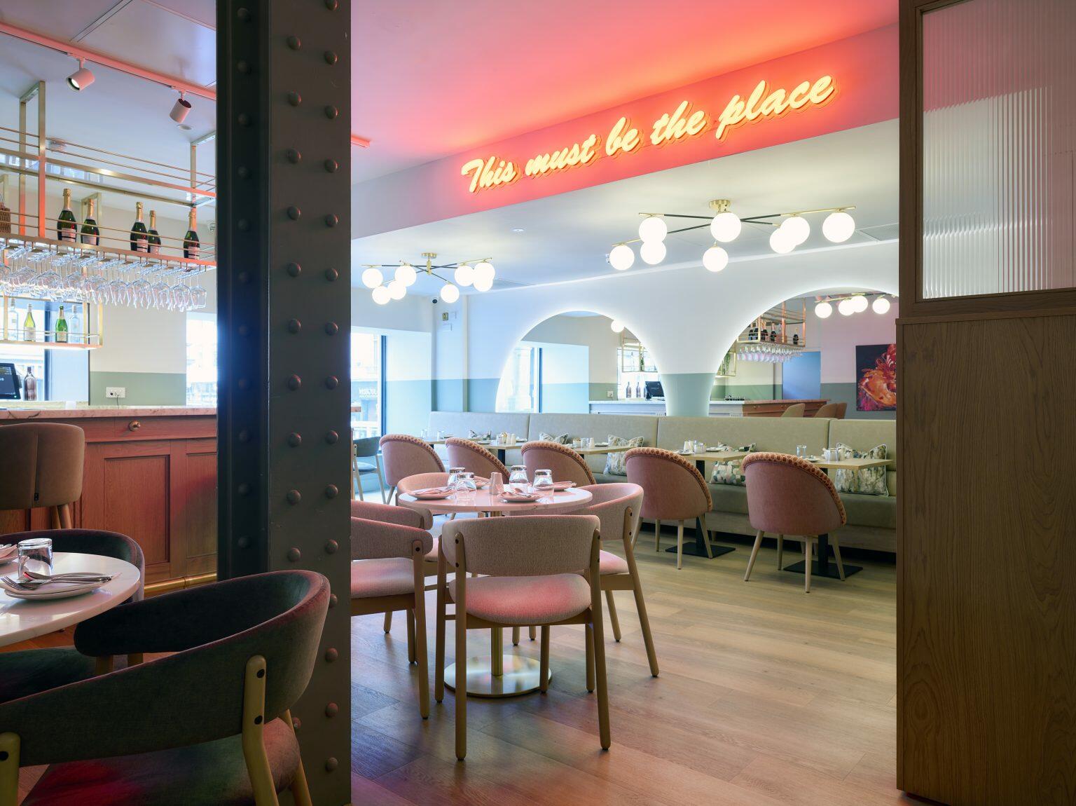 Trendy hospitality dining area with neon sign reading 'This must be the place', featuring stylish seating, marble bar top, and modern decor elements.