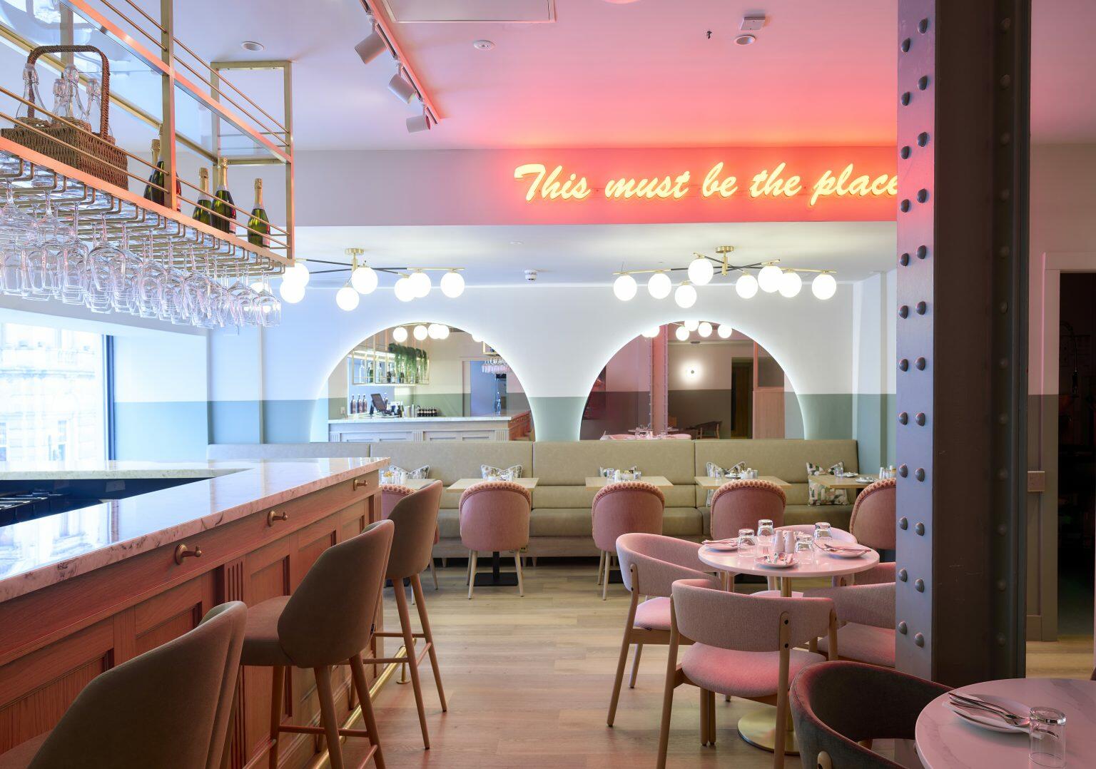 Trendy hospitality dining area with neon sign reading 'This must be the place', featuring stylish seating, marble bar top, and modern decor elements.