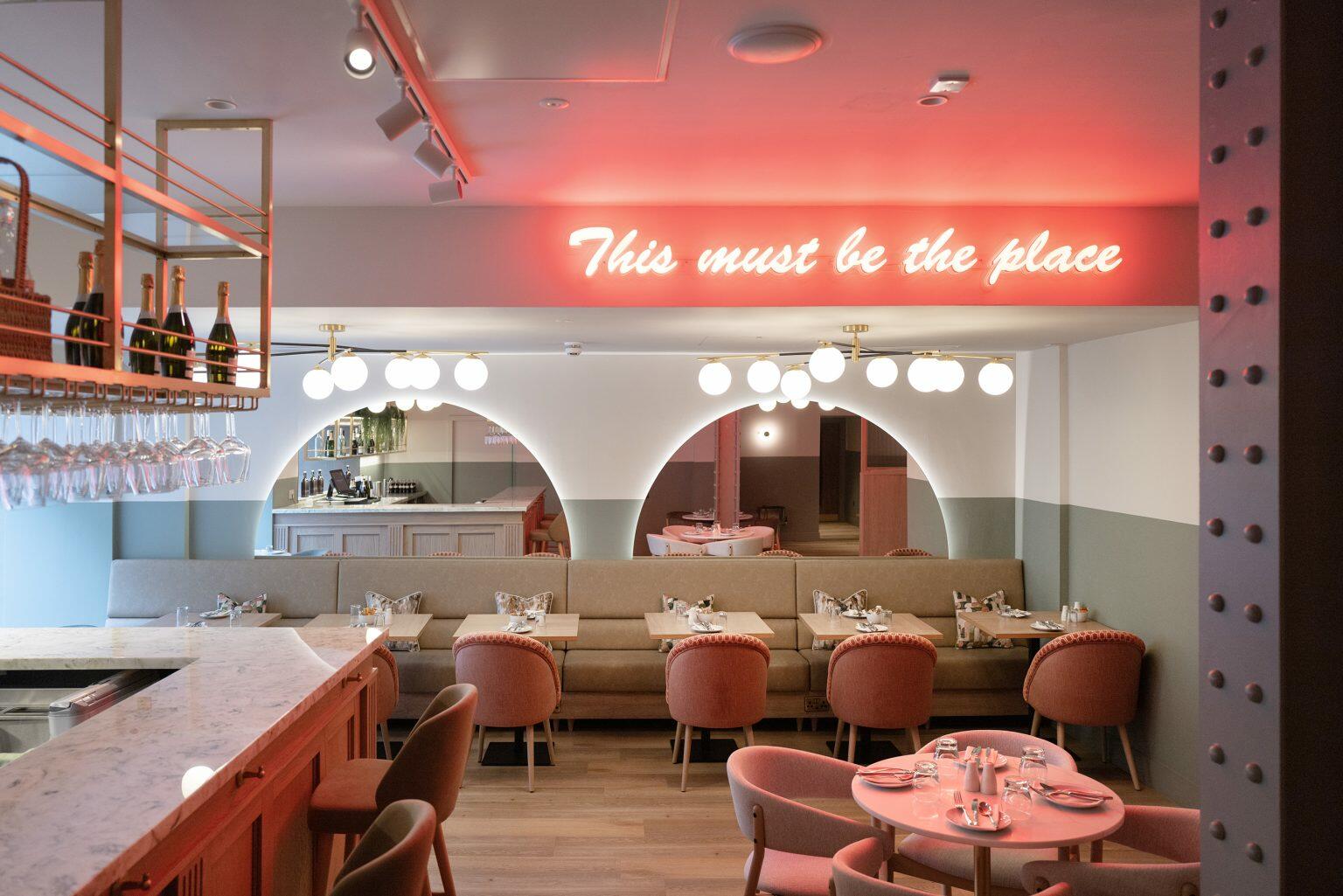 Trendy hospitality dining area with neon sign reading 'This must be the place', featuring stylish seating, marble bar top, and modern decor elements.